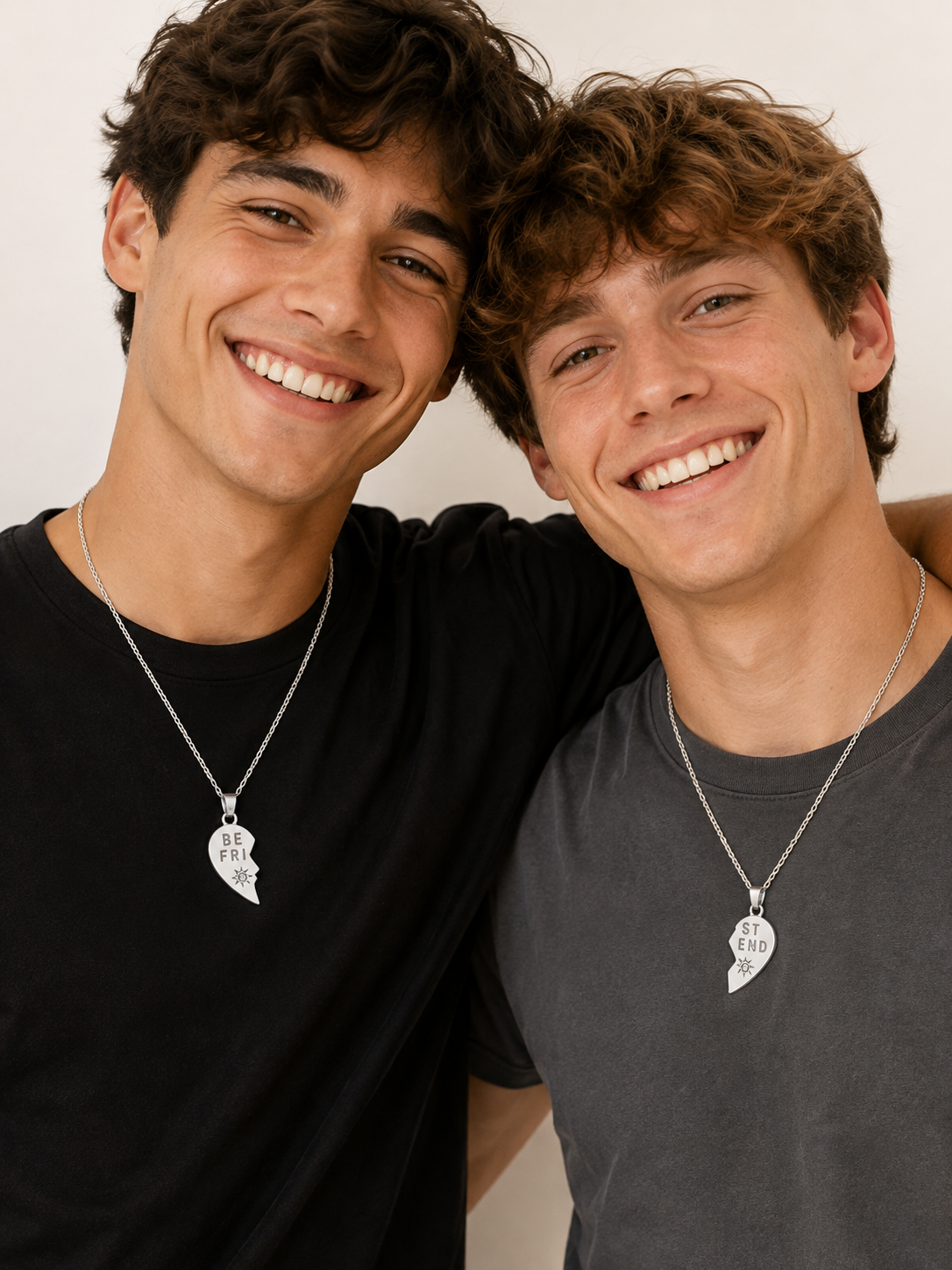 Two smiling young men standing close together, each wearing a silver necklace with a broken-heart pendant that fits together. One wears a black T-shirt and the other wears a dark gray T-shirt, against a plain light background.