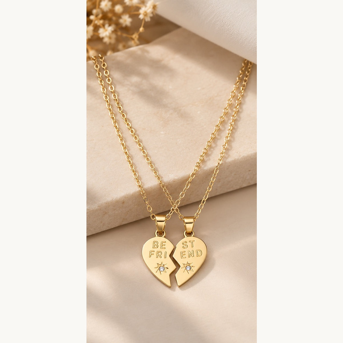 Two gold friendship necklaces with heart-shaped pendant halves engraved with 'BE FRIEND' and 'ST FRIEND', displayed on a beige stone block with dried flowers.