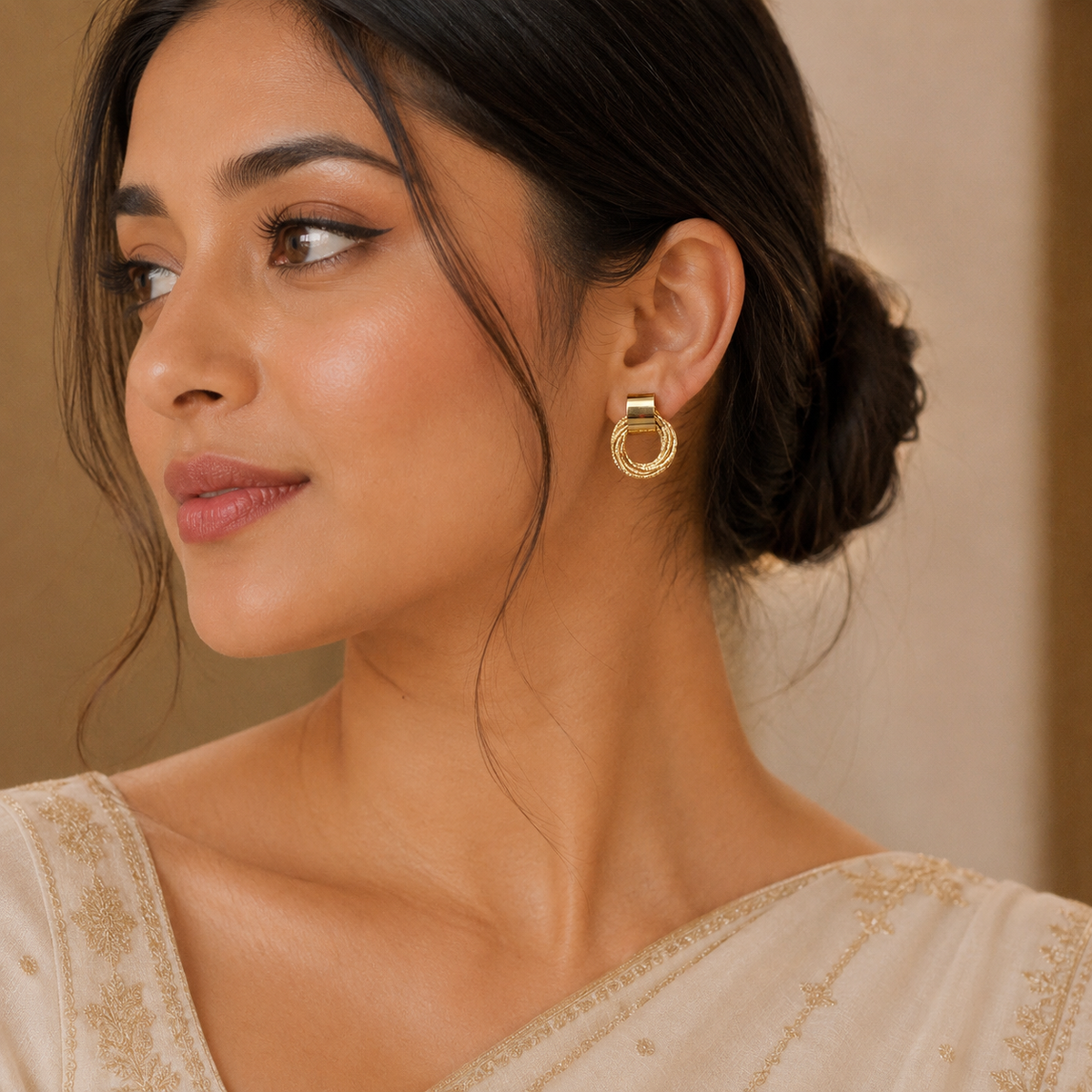 A close-up of a woman wearing a gold hoop earring with a small rectangular gold charm, shown against a warm beige background.