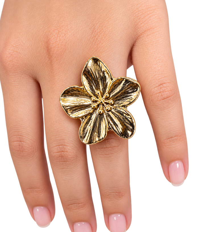 A gold flower-shaped ring worn on a hand with light pink manicured nails, set against a plain white background.
