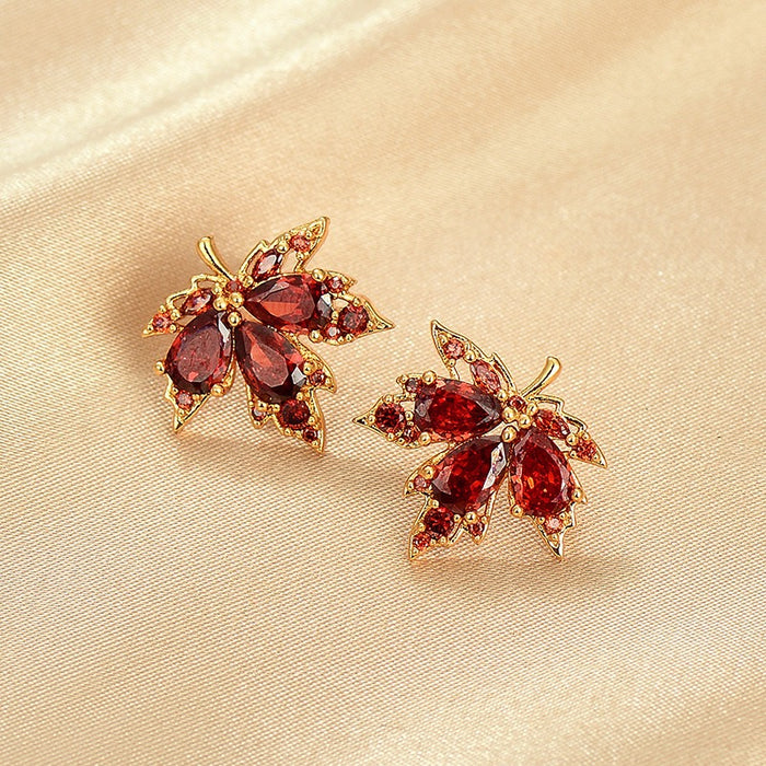 A pair of gold-toned leaf-shaped stud earrings with deep red teardrop gemstones and small clear accent stones, displayed on a soft beige satin fabric background.