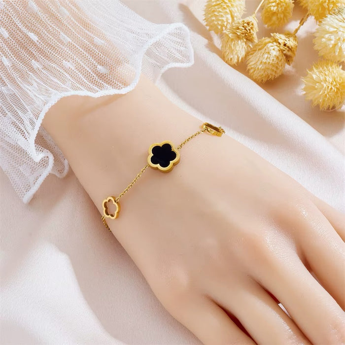 A delicate gold chain bracelet with two clover-shaped charms, one black and one gold, worn on a hand. The hand is styled with a white lace sleeve, and dried yellow flowers are in the background.