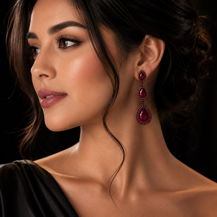 A close-up portrait of a woman wearing a long, teardrop-shaped drop earring with deep red gemstones and a textured silver-toned setting against a dark background.