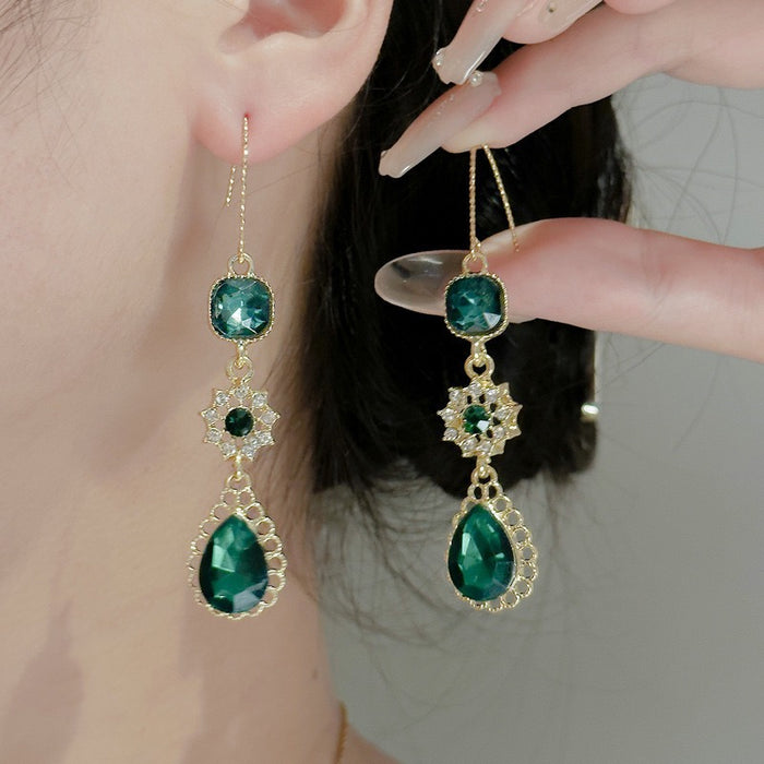 Gold-tone drop earrings with green gemstones worn by a model, with one earring hanging on the ear and the other held by a hand.