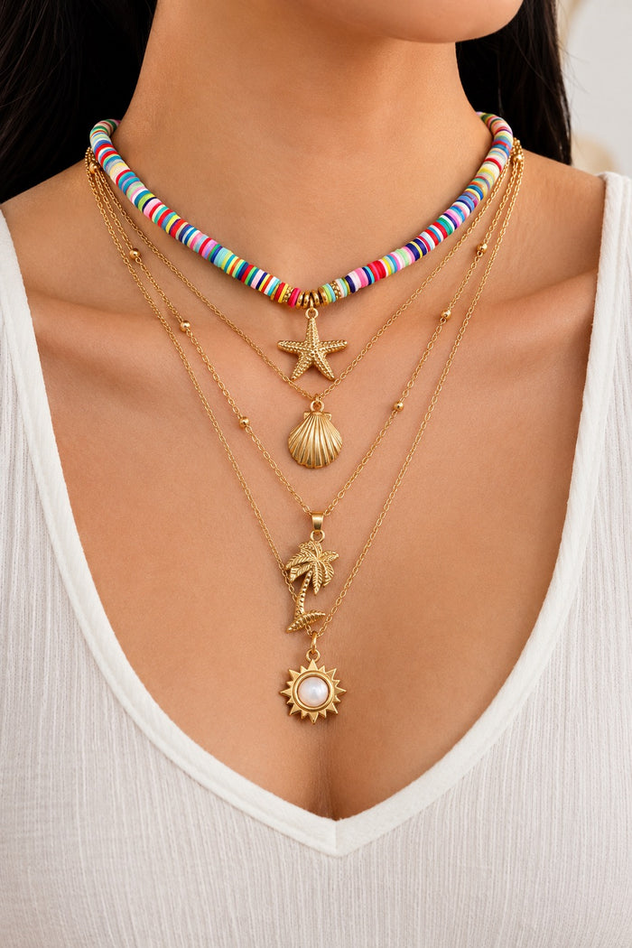 A close-up of a person wearing a layered necklace set. The set includes a colorful beaded choker, three gold-tone layered necklaces with starfish, seashell, palm tree, sun, and small bead accents, and a white V-neck top.