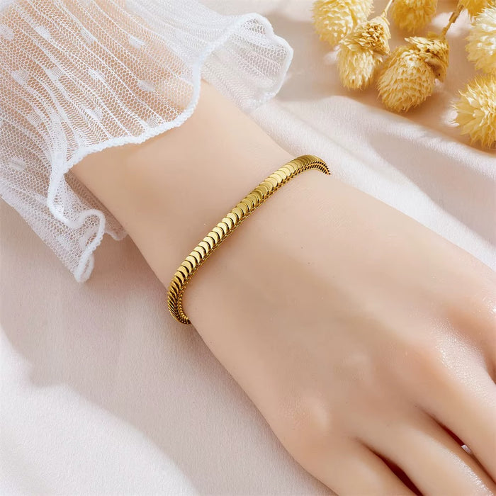 A gold-toned twisted rope bangle bracelet worn on a person's wrist, styled with a white lace sleeve and dried golden flowers on a soft white fabric background.
