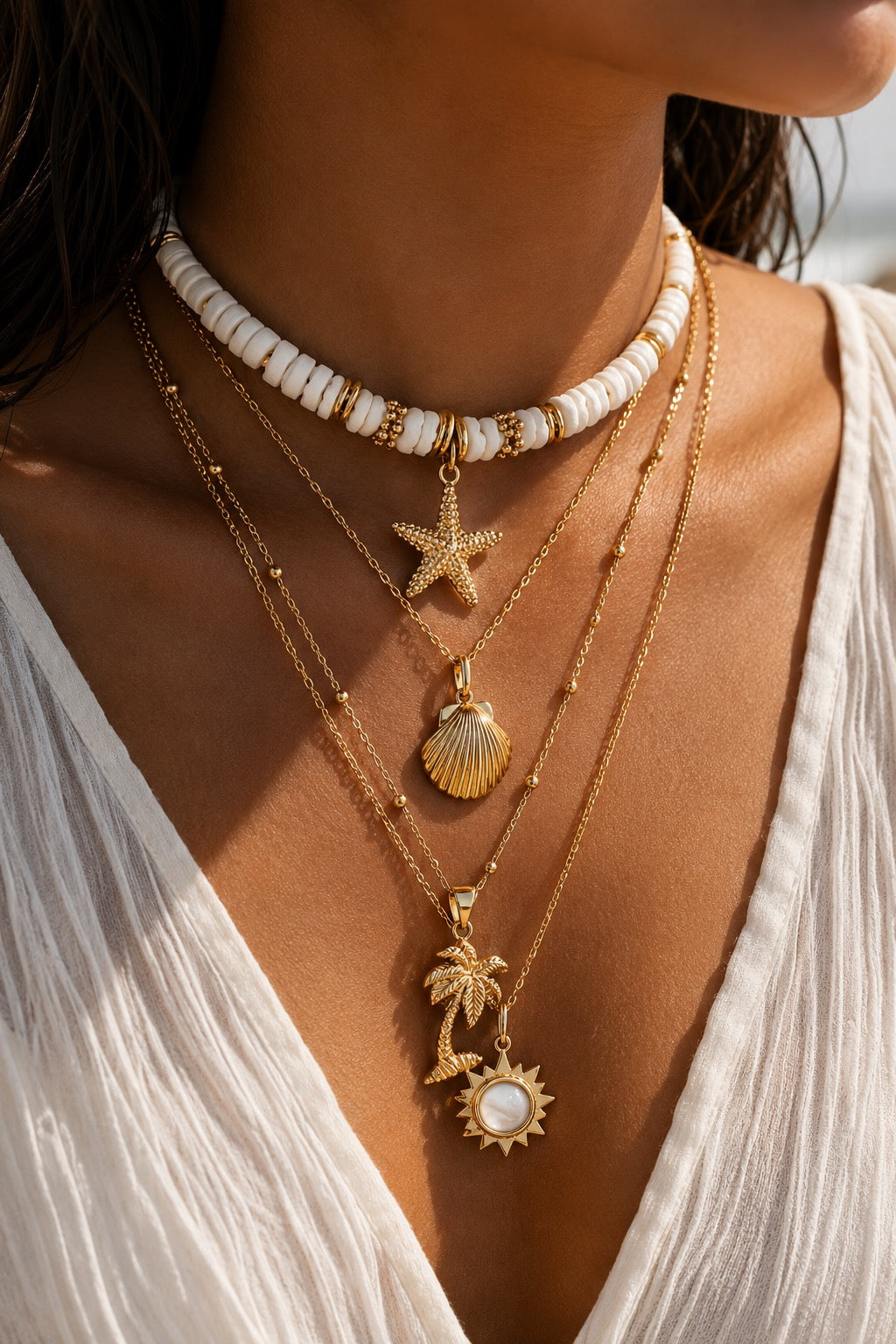 A close-up of a person wearing layered gold and white beaded necklaces, including a choker, starfish, seashell, palm tree, and sun pendant, paired with a white top.
