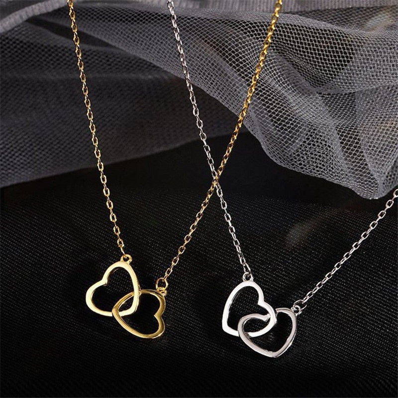 Two interlocking heart pendant necklaces displayed on black fabric with sheer gray tulle. One necklace is gold-toned and the other is silver-toned, each with a small open heart pendant on a delicate chain.