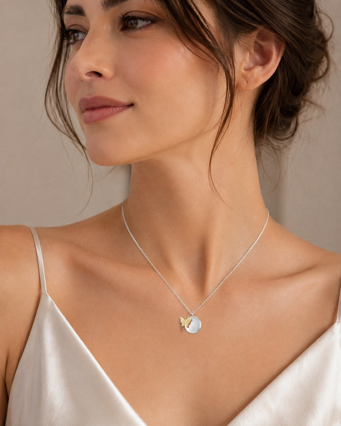 A close-up of a woman wearing a delicate silver necklace with a small white oval pendant and a tiny gold butterfly accent, paired with a white satin camisole against a neutral beige background.