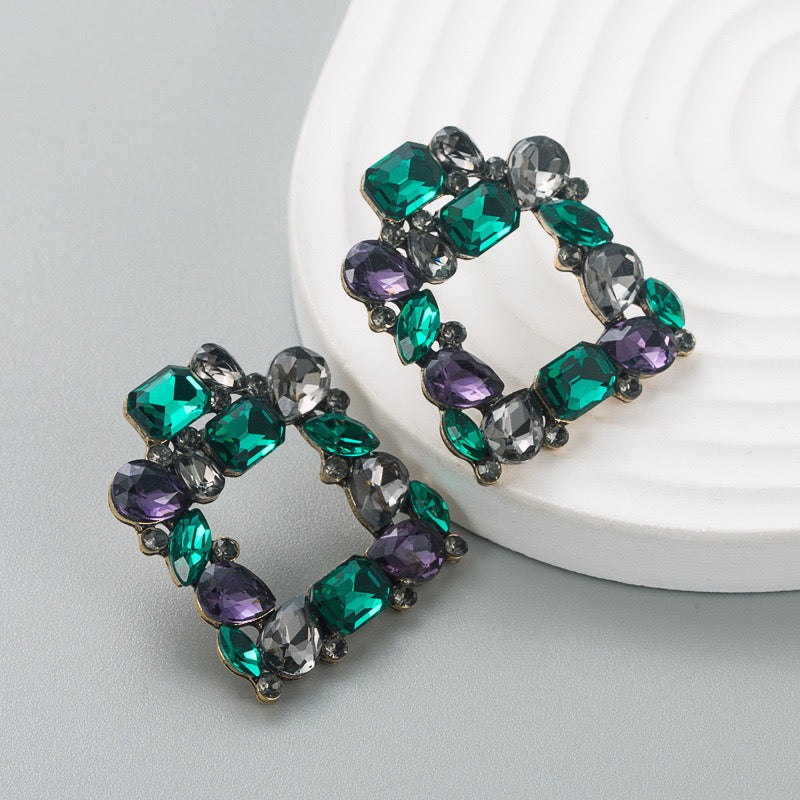 A pair of geometric hoop earrings featuring faceted green, purple, and clear rhinestones in a bronze-tone metal setting, displayed on a light gray surface next to a white textured ceramic plate.