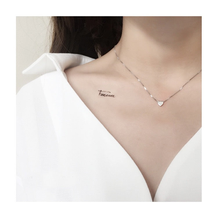 A delicate silver necklace with a small heart-shaped charm and script nameplate, shown worn on a model's collarbone over a white V-neck top.