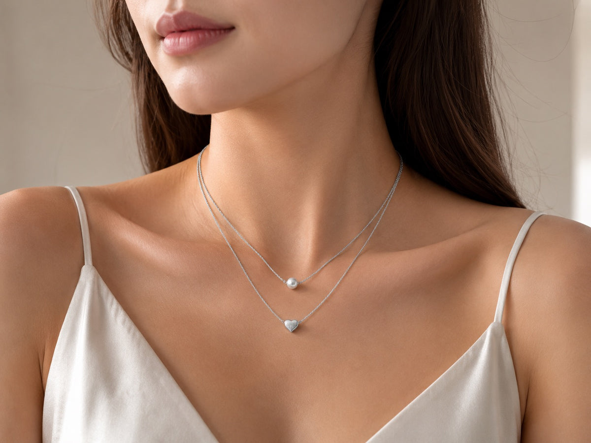 A close-up of a woman wearing a delicate double-layered silver necklace with a small pearl pendant on the top layer and a small heart-shaped pendant on the bottom layer, paired with a white camisole top.