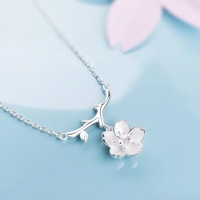 A silver necklace featuring a delicate branch pendant with a small white cherry blossom flower charm set with a pink gemstone, displayed on a soft light blue background.