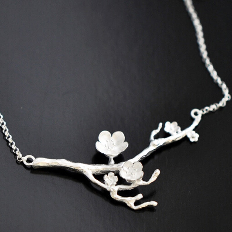 A silver branch pendant necklace with three textured cherry blossom flowers, displayed on a dark black background.