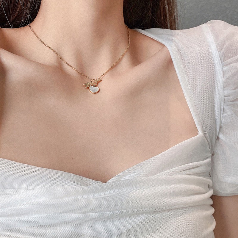 A close-up of a model wearing a delicate gold angel necklace with a small white heart pendant, styled with a white sheer top.