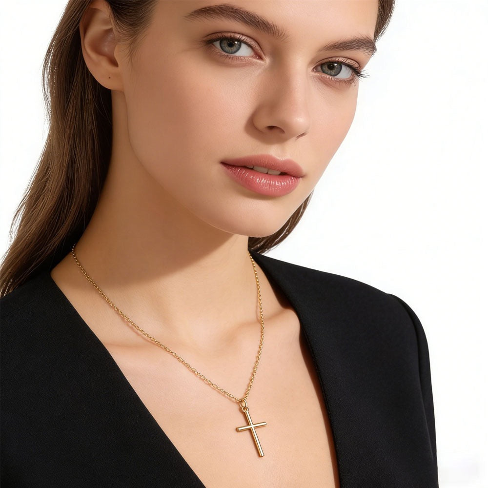 A close-up of a woman wearing a delicate gold cross pendant necklace on a fine chain, paired with a black top against a clean white background.