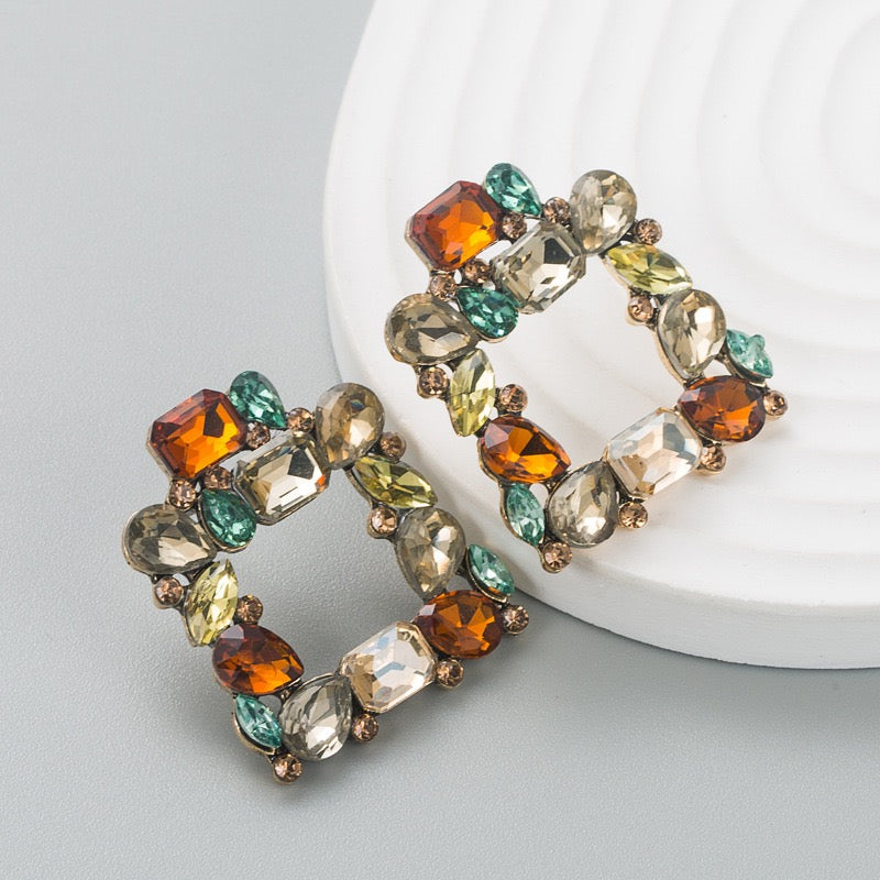 A pair of square hoop earrings adorned with faceted rhinestones in amber orange, emerald green, pale yellow, and clear crystal, set in a bronze-toned metal, displayed on a white textured circular plate against a light gray background.