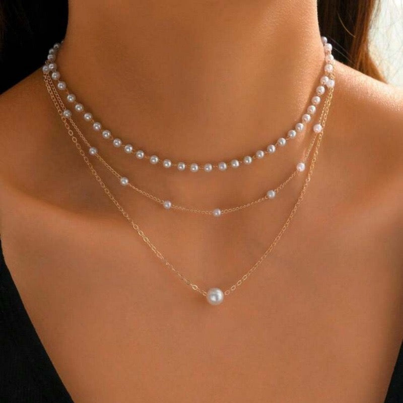 A close-up of a model's neck and upper chest wearing a layered necklace set. The set includes a short pearl strand, a longer gold chain with small pearl beads, and a longer gold chain with a single larger pearl pendant.