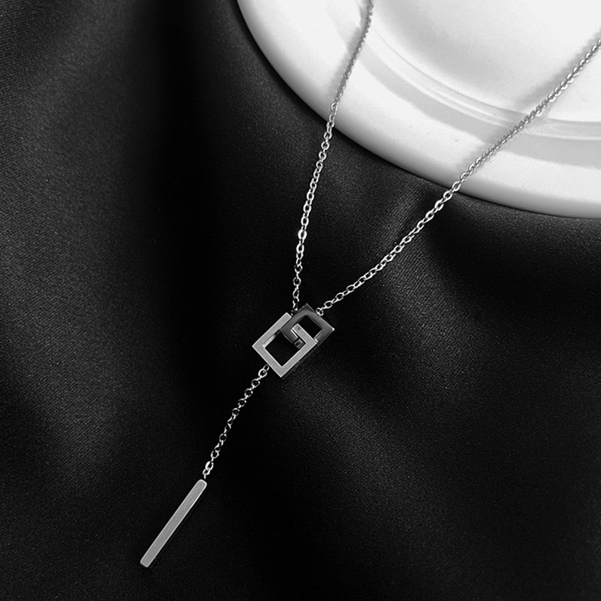 A silver-toned geometric lariat necklace displayed on a black satin fabric bust form with a white ceramic neck piece visible at the top. The necklace features a fine chain, an open square pendant with a stylized interlocking design, and a long flat bar dangling from the chain.