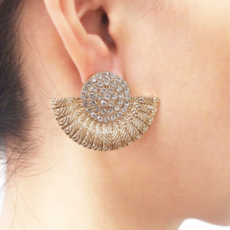 A close-up of a model's ear wearing a gold crescent-shaped stud earring with a textured leaf pattern and a round crystal-encrusted center.