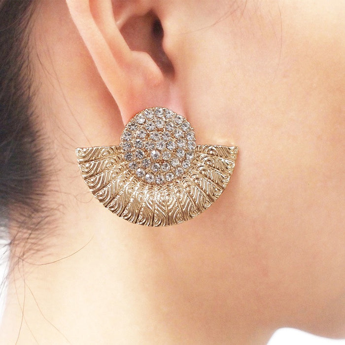 A close-up of a model's ear wearing a gold crescent-shaped stud earring with a textured leaf pattern and a round crystal-encrusted center.
