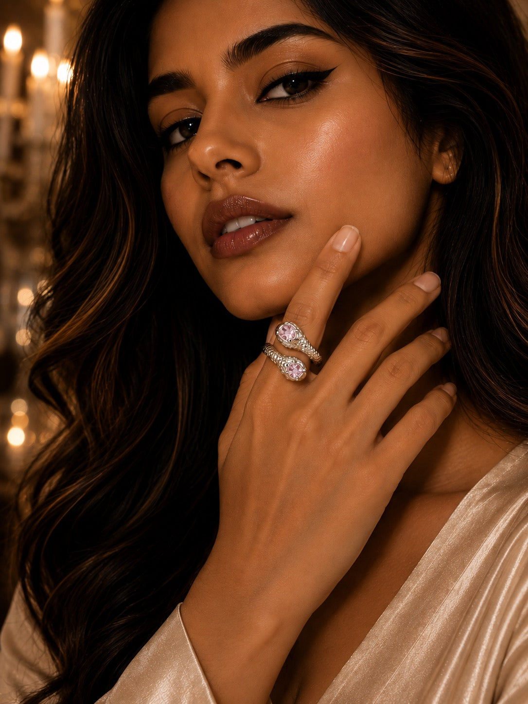 A close-up lifestyle shot of a woman wearing a distinctive double-helix ring with sparkling clear gemstones. The warm, atmospheric background features soft golden bokeh lights, and she is wearing a champagne satin robe.