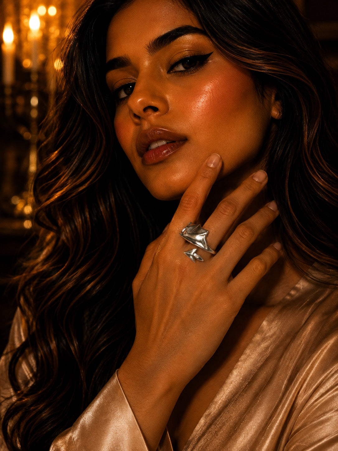 A close-up lifestyle shot of a woman with long wavy brown hair wearing a large, chunky silver ring with a starburst or flower-like design on her finger. She is wearing a champagne-colored satin robe against a warm, softly blurred background with golden bokeh lights.