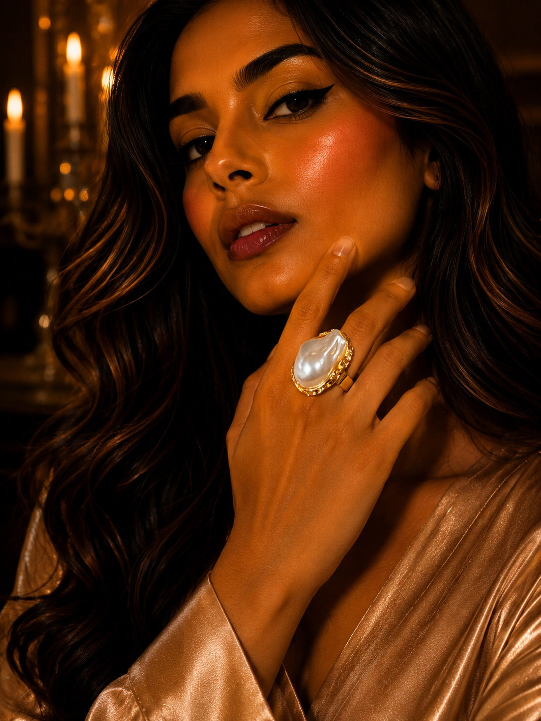 A close-up lifestyle shot of a woman with long wavy dark hair wearing a large statement ring with a white pearl-like center and gold setting, while wearing a shiny champagne satin robe.
