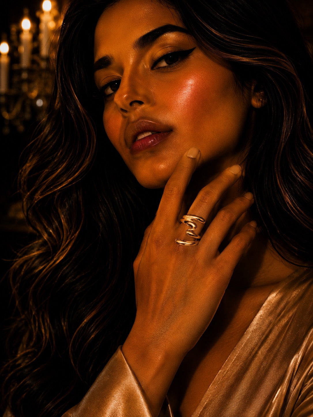 A close-up portrait of a woman with long wavy dark hair, wearing multiple stacked silver rings on one finger, in warm candlelit lighting.