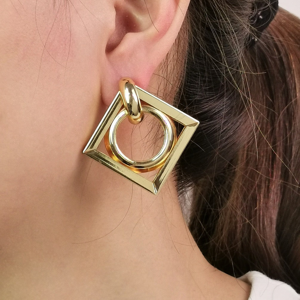 A close-up of a person's ear wearing a large gold geometric earring with a square frame and circular ring detail.