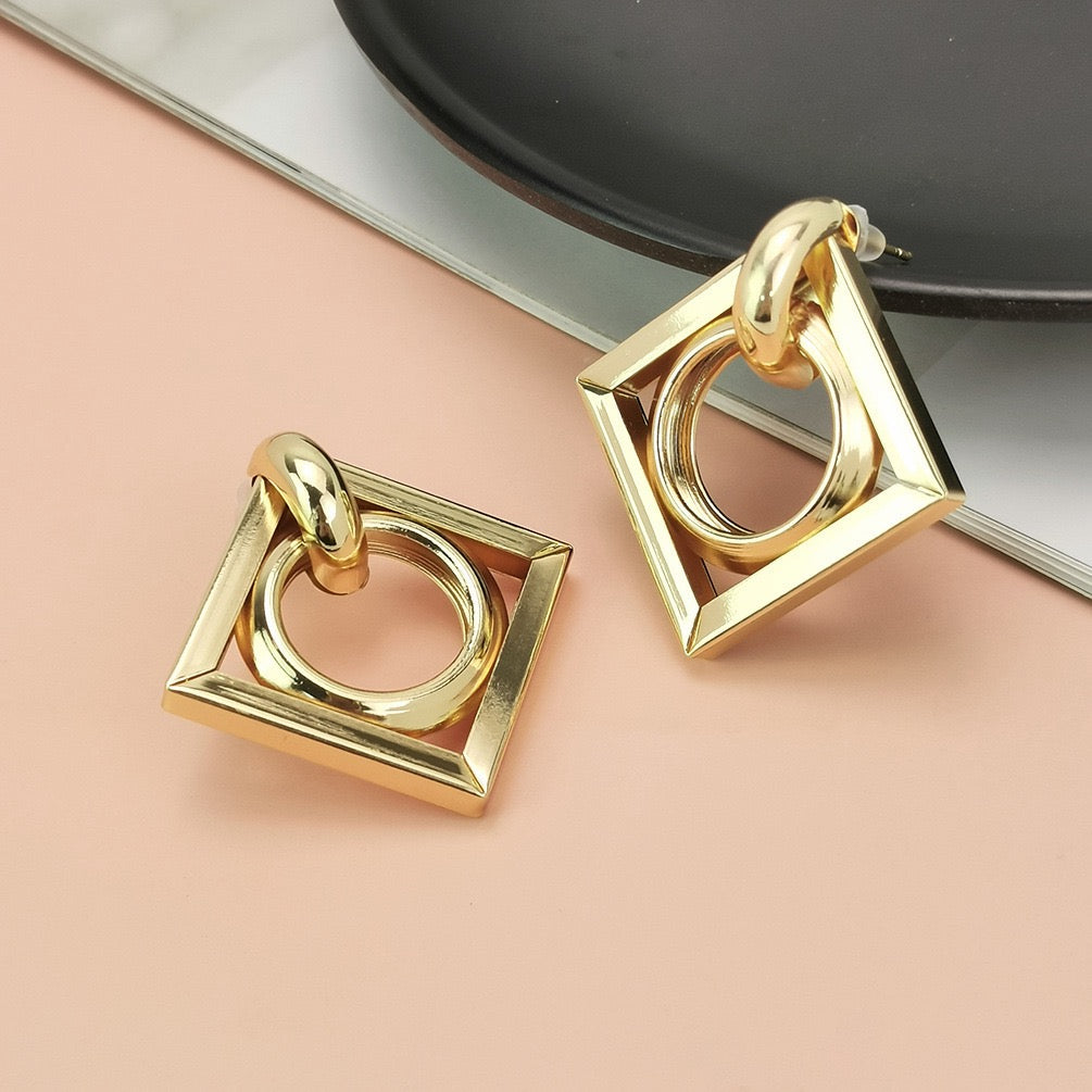 A pair of gold geometric square drop earrings with a central circular ring, displayed on a peach-colored surface with a black plate in the background.