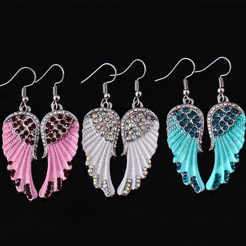 Three pairs of dangling angel wing earrings on a black background. The left pair is pink with dark red rhinestones, the center pair is white with iridescent rhinestones, and the right pair is turquoise with dark blue rhinestones.