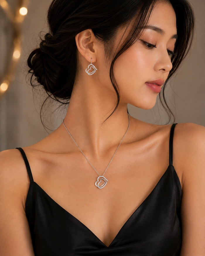 A woman wearing a delicate silver necklace and matching heart-shaped stud earring, styled with a black satin camisole against a softly blurred warm-toned background.