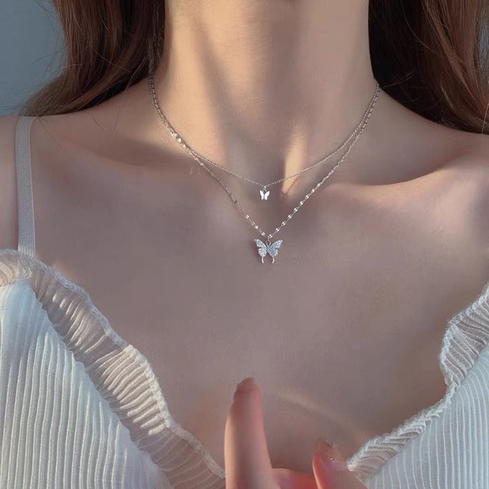 A close-up of a model's neck and chest wearing a delicate layered silver necklace with two small butterfly pendants, styled with a white ruffled top.