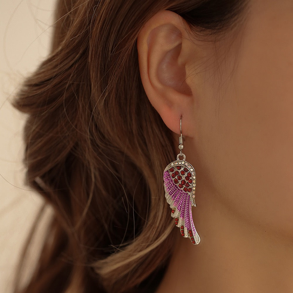 A close-up of a woman's ear wearing a dangling wing-shaped earring. The earring has a silver-toned metal frame, clear rhinestone trim, and pink and red beadwork.