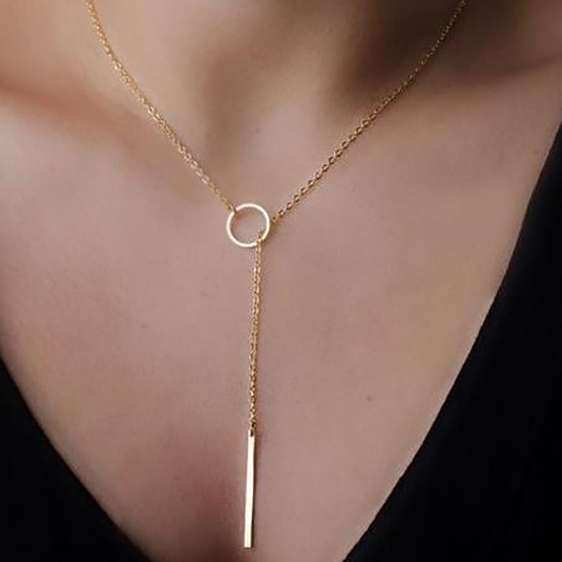 A gold lariat-style necklace with a circular ring at the center and a hanging bar pendant, shown worn on a model's neck over a black top.