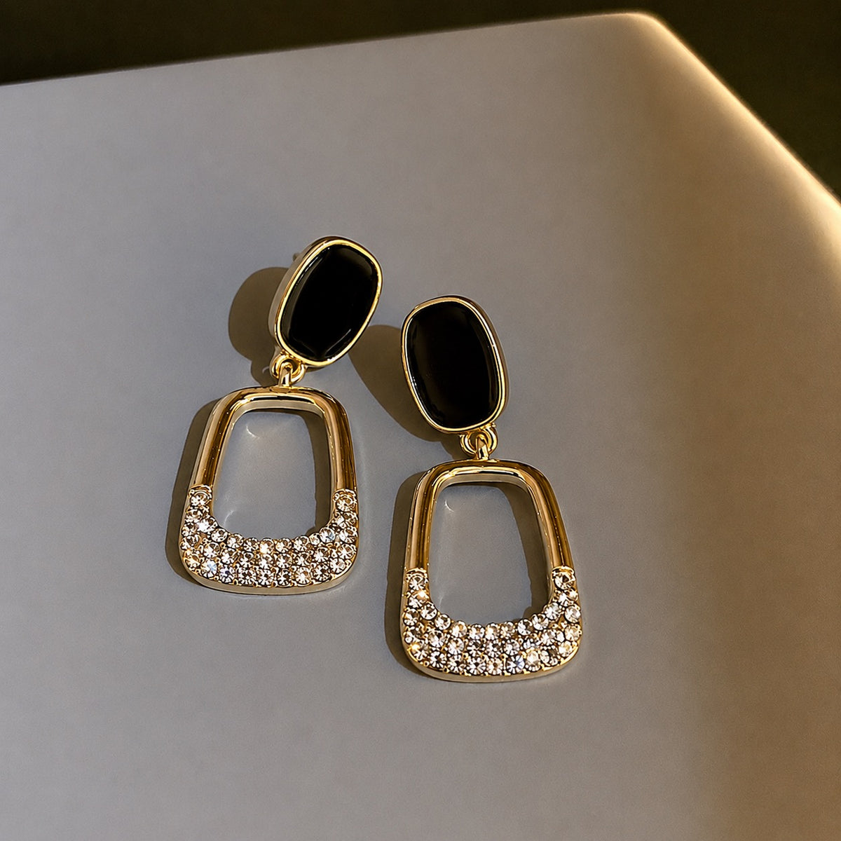 A pair of gold-tone statement dangle earrings with black oval studs and open arch-shaped drops paved with clear rhinestones, resting on a light gray surface.
