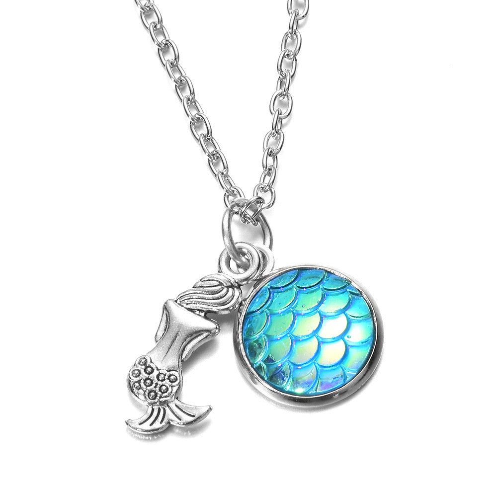 A silver-tone chain necklace featuring two charms: a small mermaid tail charm and a round iridescent blue-green scale charm, shown on a plain white background.