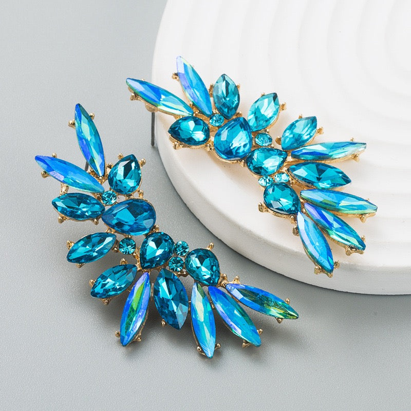 A pair of gold-tone statement earrings featuring iridescent blue marquise and oval rhinestones, displayed on a light gray surface with a white textured circular prop.