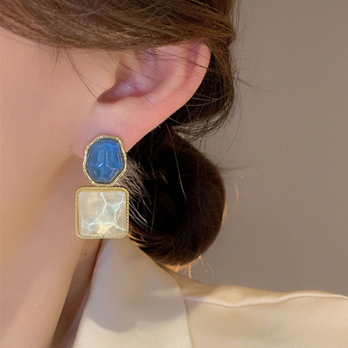 A close-up of a model's ear wearing a two-step drop earring. The top step features a faceted blue stone in a gold bezel, and the bottom step is a square milky white stone in a gold bezel.