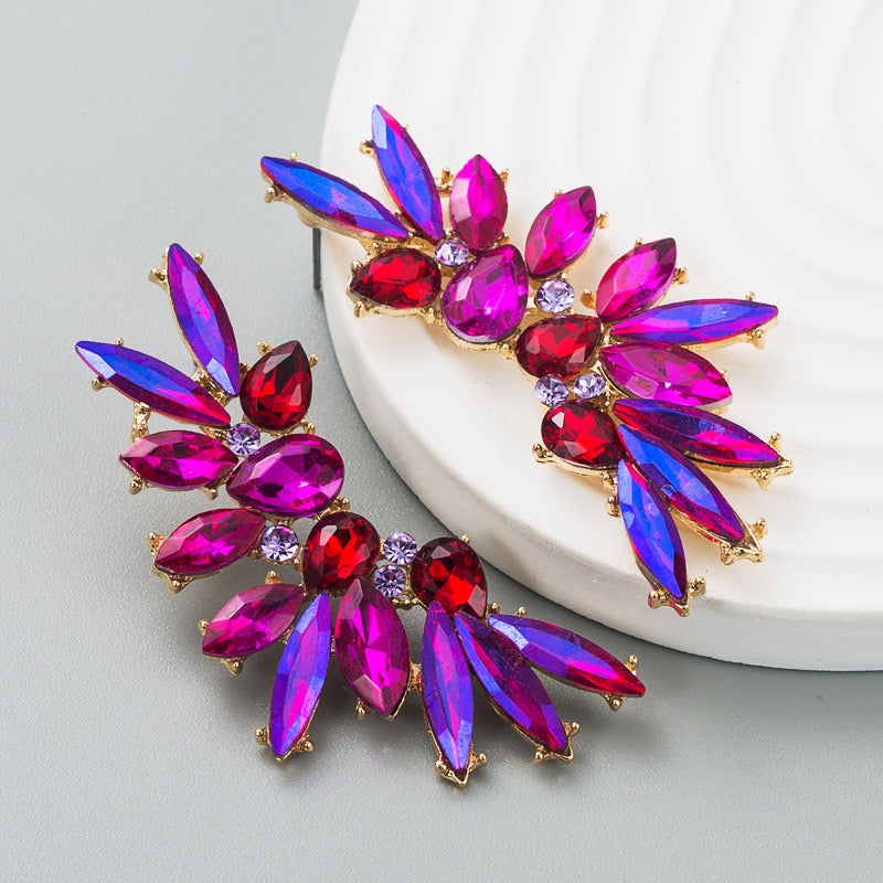 A pair of gold-tone floral brooch pins featuring iridescent magenta and purple marquise crystals with small red teardrop crystals and clear accent rhinestones, displayed on a white ceramic plate against a light gray background.