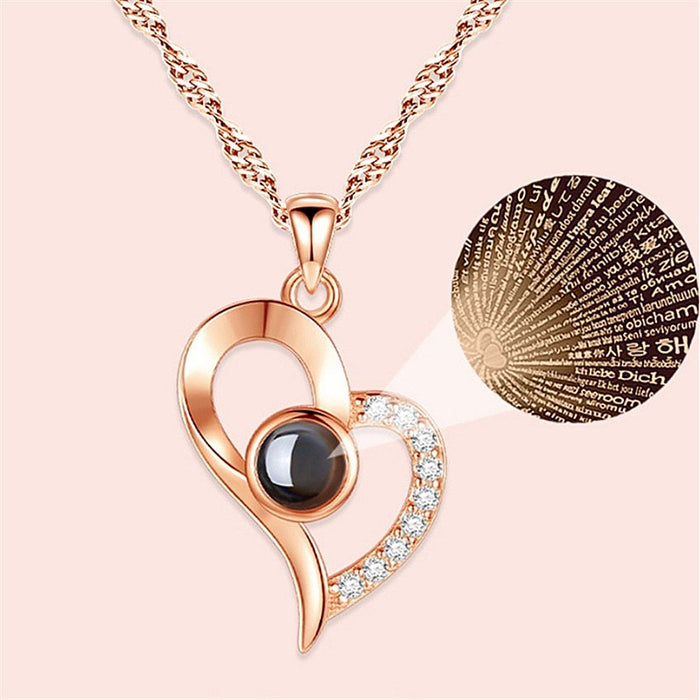 Rose gold heart-shaped pendant necklace with a central black gemstone and a side of clear crystals, shown on a light pink background with a circular detail inset of engraved text.