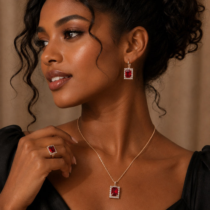 A woman wearing a matching gold jewelry set with rectangular red gemstones, including a pendant necklace, ring, and drop earring, shown against a warm beige background.