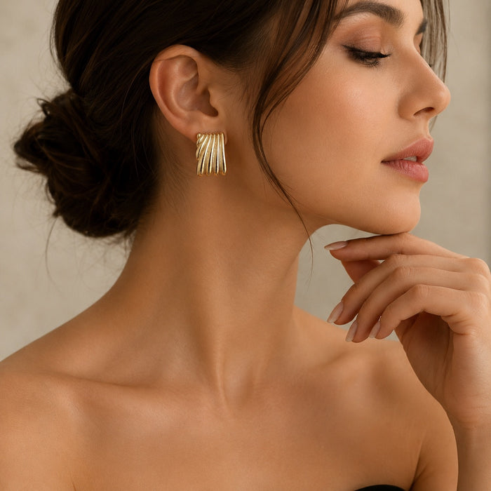 A close-up of a woman wearing a pair of gold geometric drop earrings with multiple parallel bar details, set against a soft beige background.