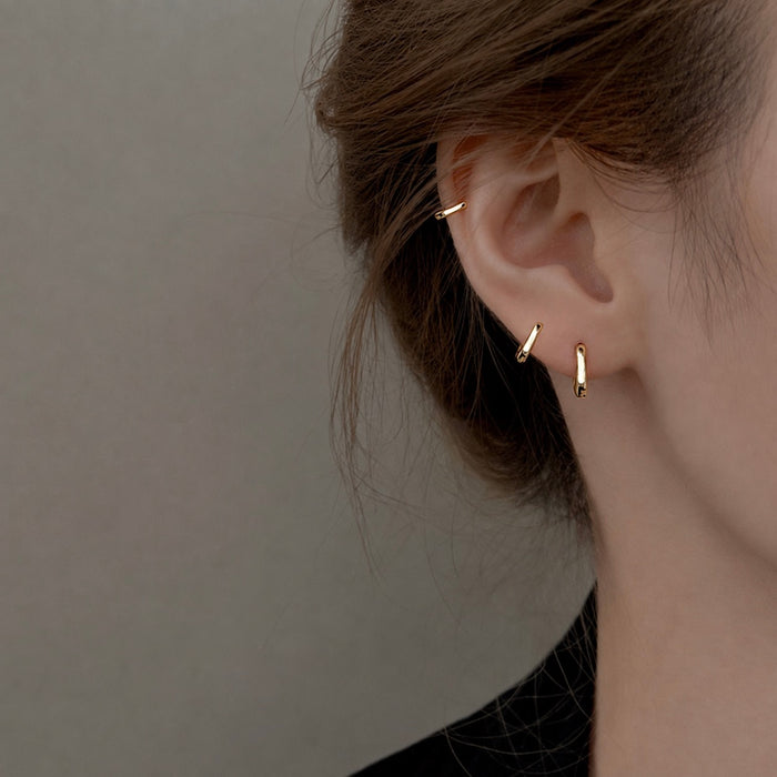 A close-up of a person's ear wearing three delicate gold earrings: a small hoop in the lobe, a tiny curved bar above, and a small vertical bar below. The background is a soft, neutral gray.