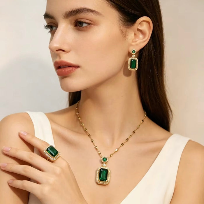 A woman models a matching gold jewelry set featuring a necklace, ring, and drop earring, each with a rectangular emerald-cut green gemstone surrounded by small clear stones.
