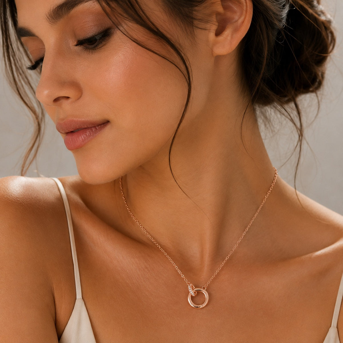 A close-up lifestyle shot of a woman wearing a delicate rose gold necklace with a minimalist open circle pendant, styled with a cream camisole against a soft neutral background.