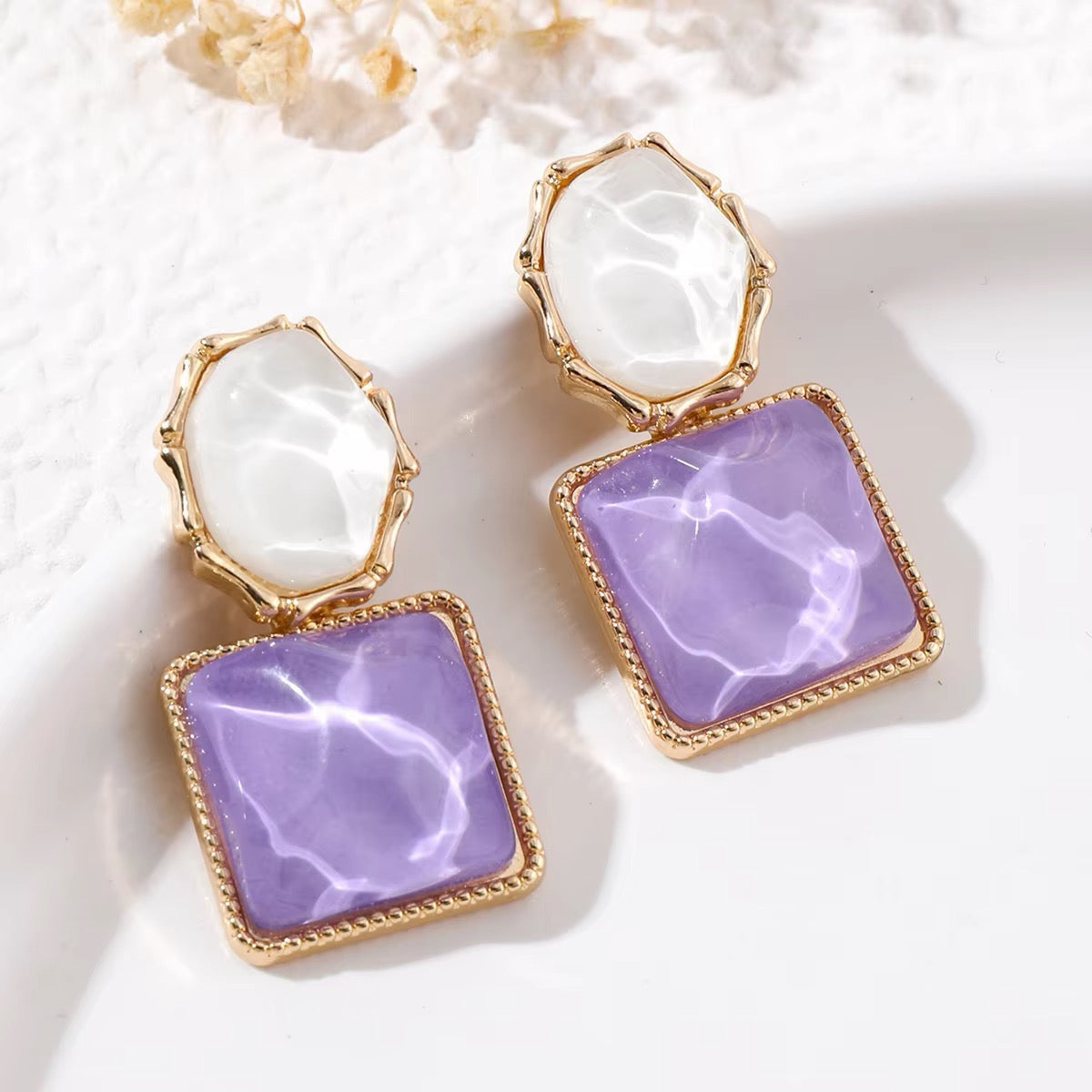 A pair of gold-tone drop earrings featuring a large square lavender-purple stone on the bottom and an oval white stone on the top, displayed on a white textured surface with soft shadows.