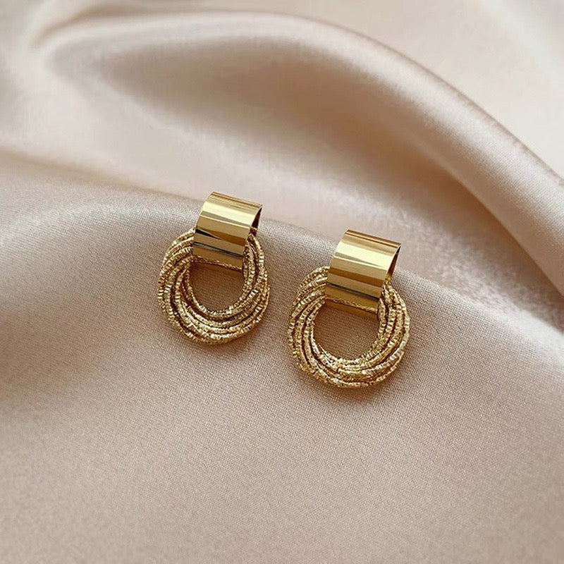 A pair of gold hoop earrings with textured, multi-strand beaded rings and polished rectangular posts, displayed on a draped beige satin fabric.