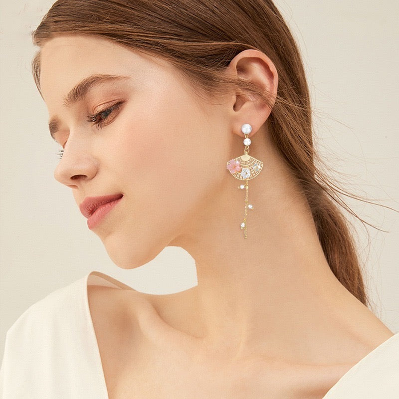 A woman wearing a dangling earring with a gold-tone fan-shaped setting, white pearl-like beads, and iridescent pastel accents, shown in a close-up portrait against a soft light beige background.